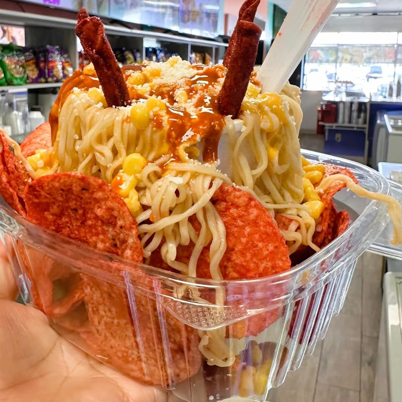 A hand holds a clear plastic container filled with ramen noodles, corn, red chips, and sauce.