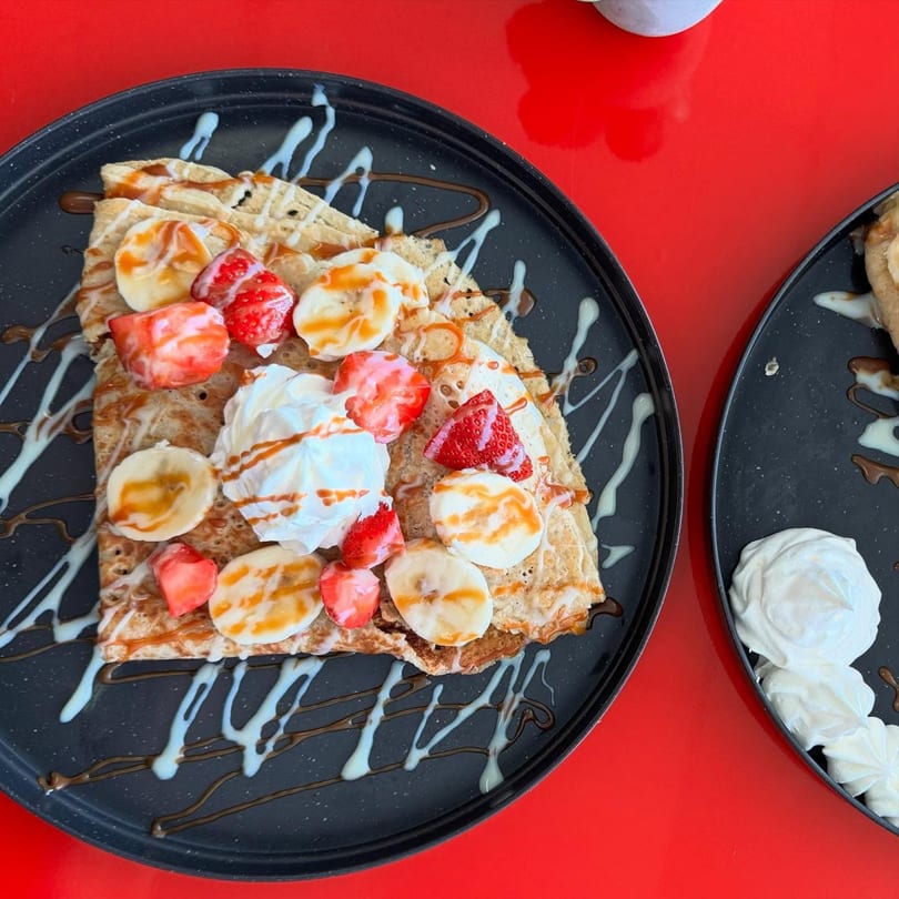 A crepe topped with strawberries, bananas, whipped cream, and caramel drizzle on a black plate.