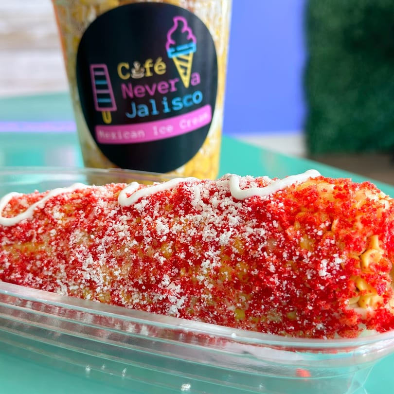 Elote with red chili powder, white cheese, and cream, with a Jalisco Mexican Ice Cream cup in the background.