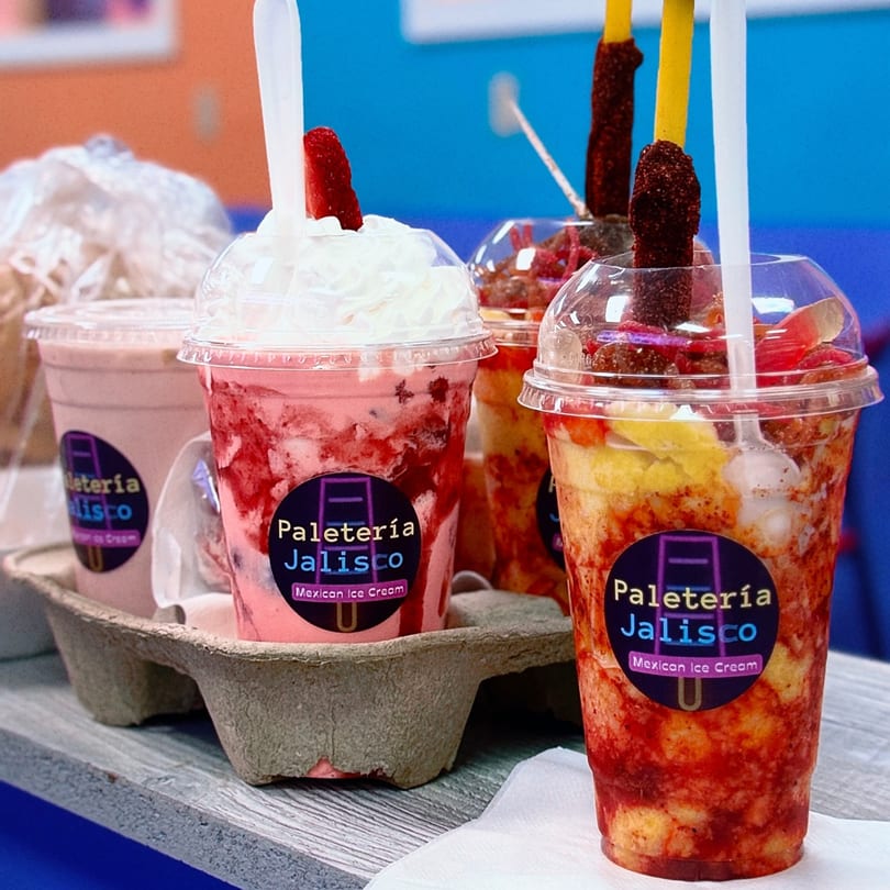 Four colorful Paletería Jalisco Mexican Ice Cream drinks, two with fruit and candy, one with whipped cream.